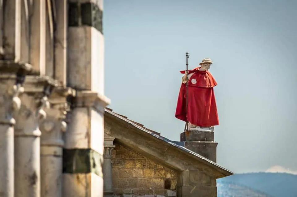 Statua di un pellegrino con mantello rosso, cappello e bastone, sopra un tetto accanto a una facciata in pietra con colonnine