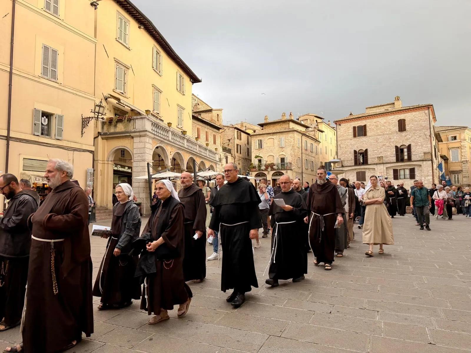 Corteo di frati in tonaca marrone e suore che attraversa una piazza storica circondata da edifici in pietra, con altre persone che seguono la processione.