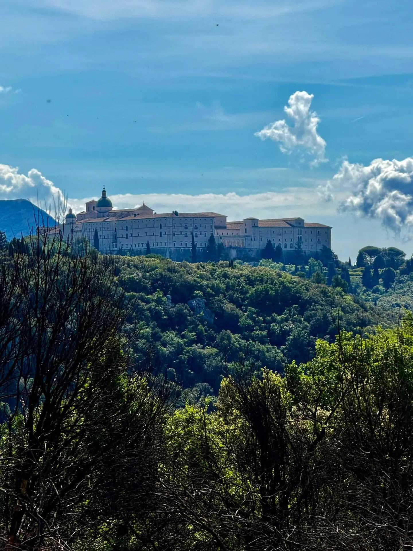 Grande complesso monastico in pietra chiara sulla sommità di una collina coperta da boschi, sotto un cielo con nuvole sparse.