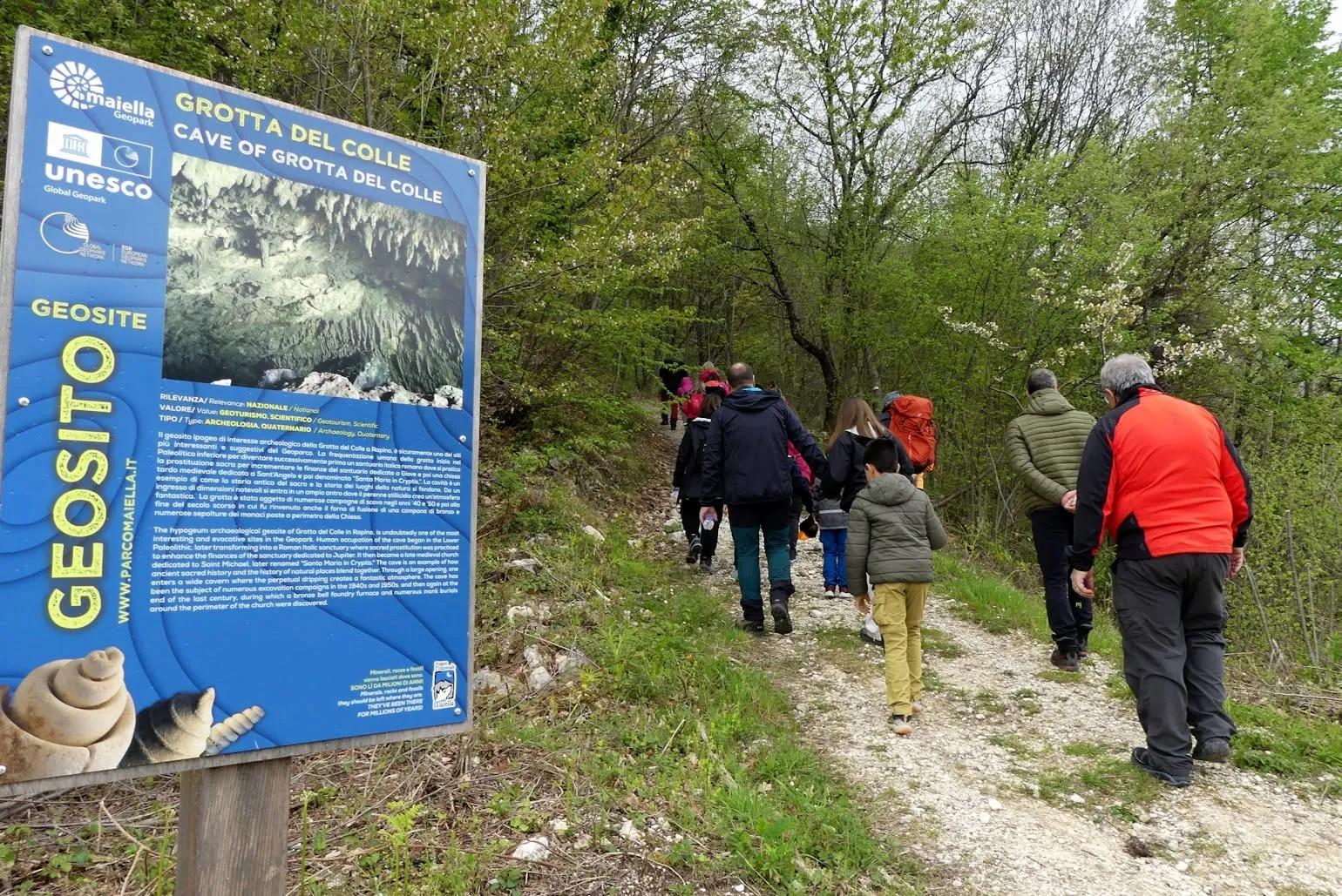 Gruppo di escursionisti, anche con bambini, cammina su un sentiero nel bosco accanto al pannello del geosito UNESCO Grotta del Colle