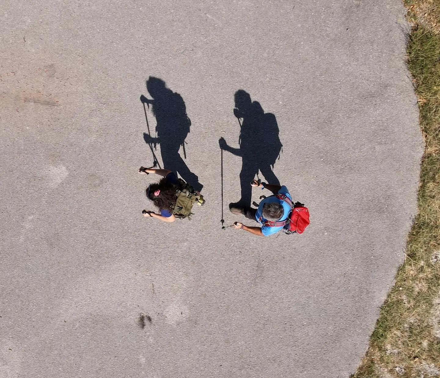 Veduta aerea di due escursionisti con zaino e bastoncini che camminano su una strada asfaltata, con lunghe ombre sul fondo