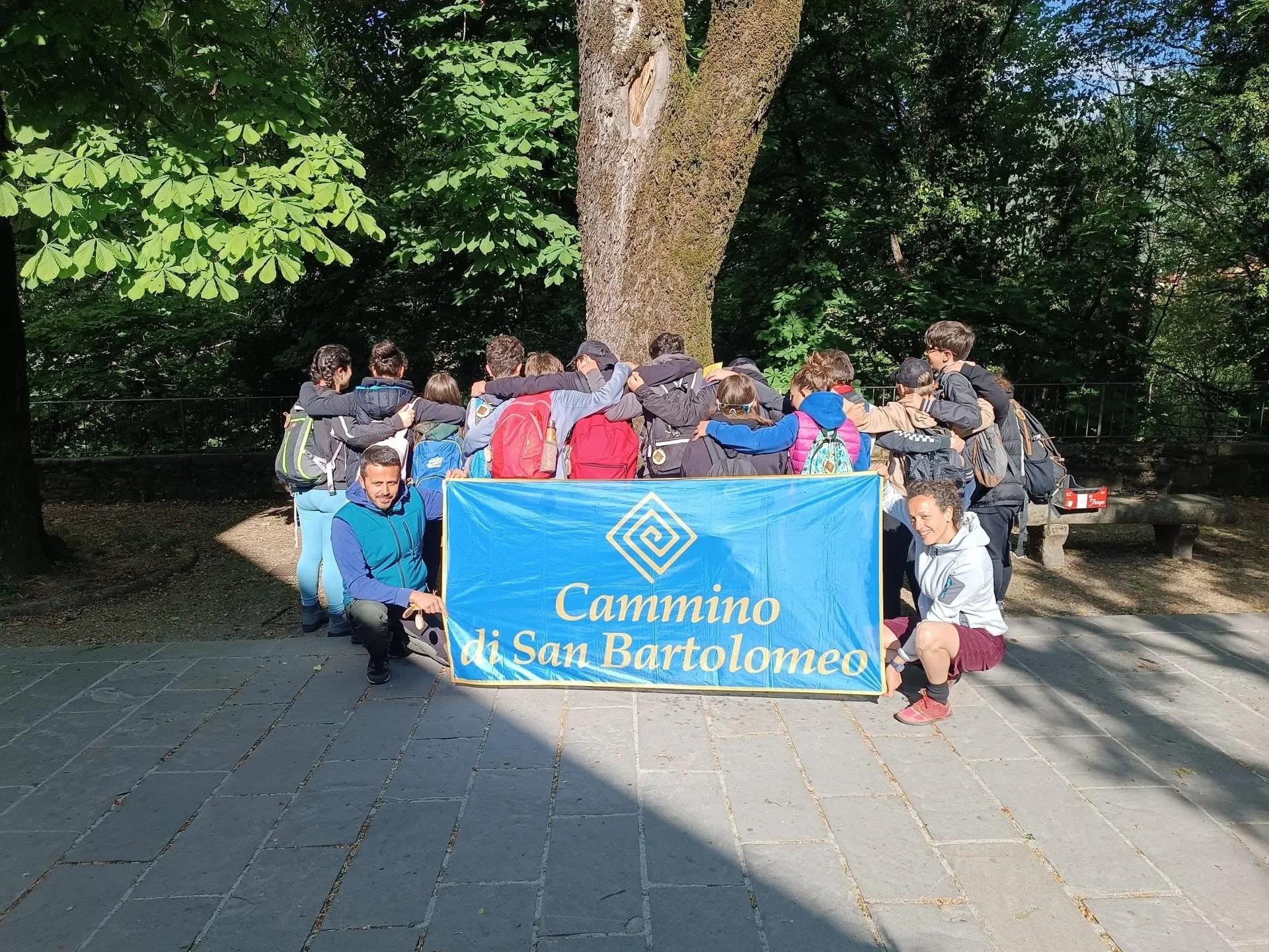 Gruppo di camminatori raccolti in cerchio sotto un grande albero, con lo striscione azzurro del Cammino di San Bartolomeo