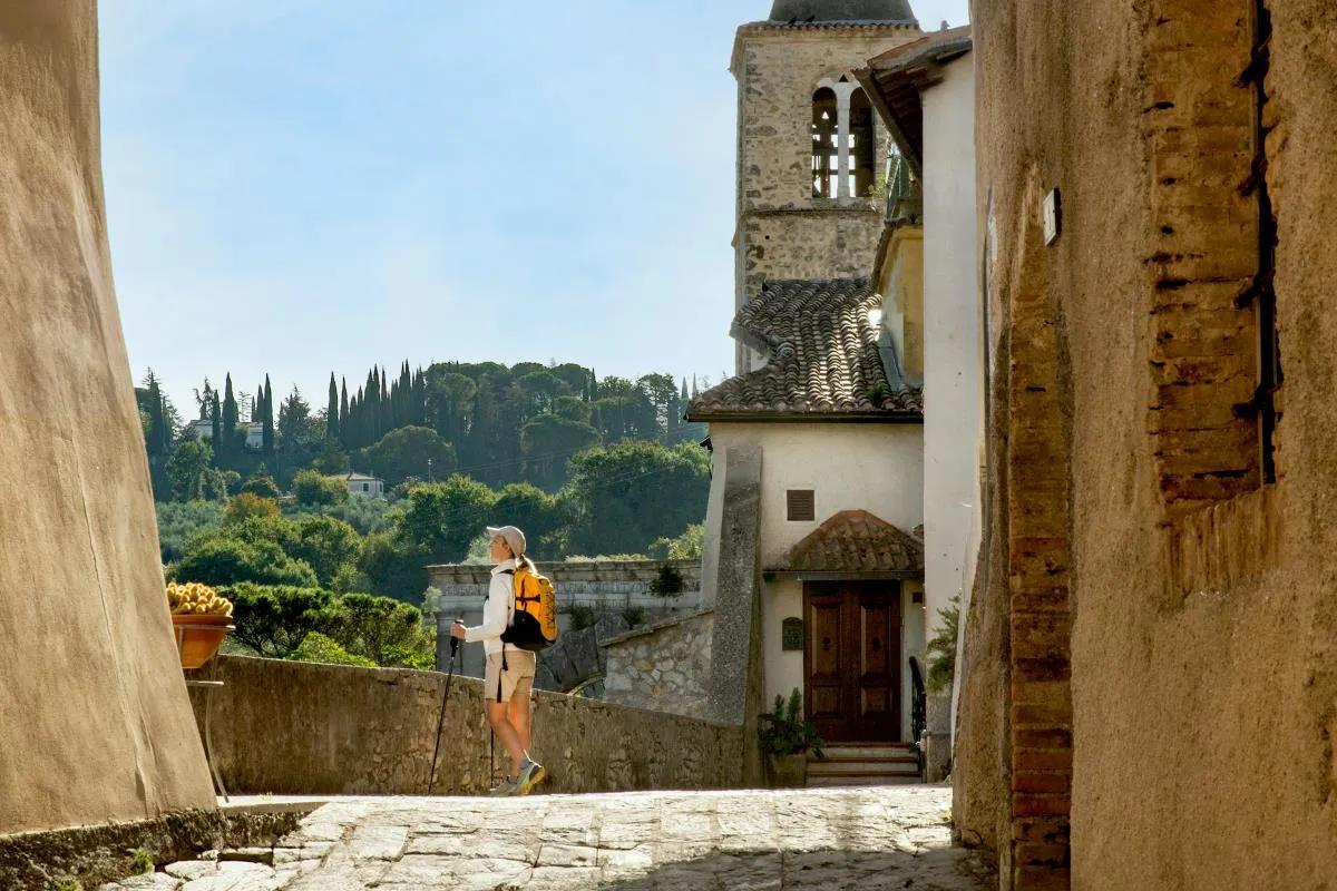 Escursionista con zaino arancione cammina lungo un vicolo in pietra di un borgo, con cipressi e colline sullo sfondo