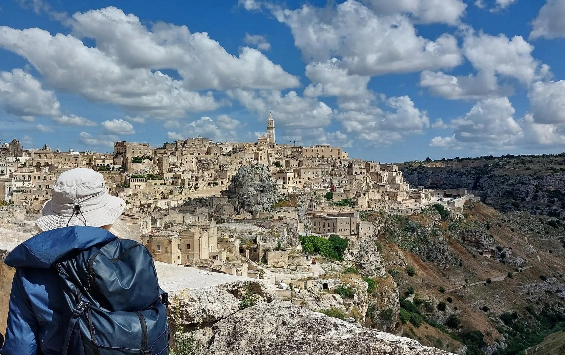 Veduta dei Sassi di Matera, con le case in pietra, il campanile in alto e la gravina sulla destra; in primo piano un escursionista di spalle con cappello bianco e zaino scuro, sotto un cielo azzurro con nuvole.