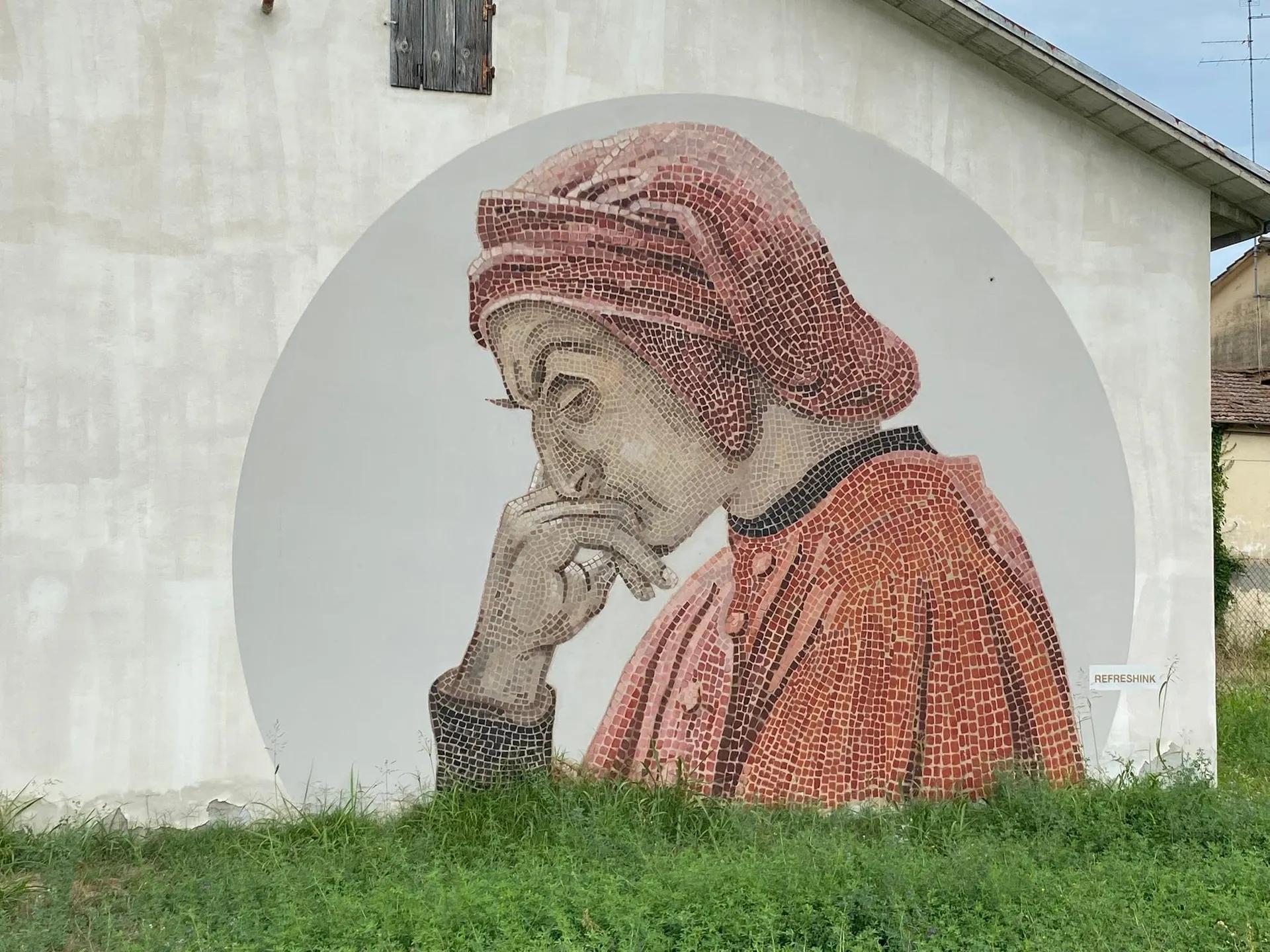 Murale a mosaico di Dante di profilo con cappuccio rosso e mano al mento, su una parete con erba in primo piano
