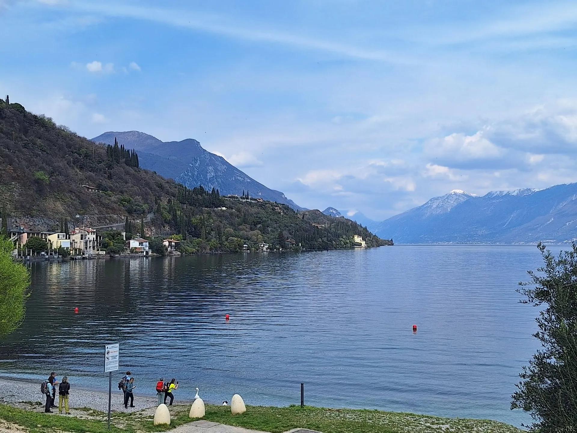 Un gruppo di escursionisti con lo zaino sulla riva ghiaiosa di un lago, con un versante boscoso e qualche casa sull’acqua e montagne con la neve in cima sullo sfondo.