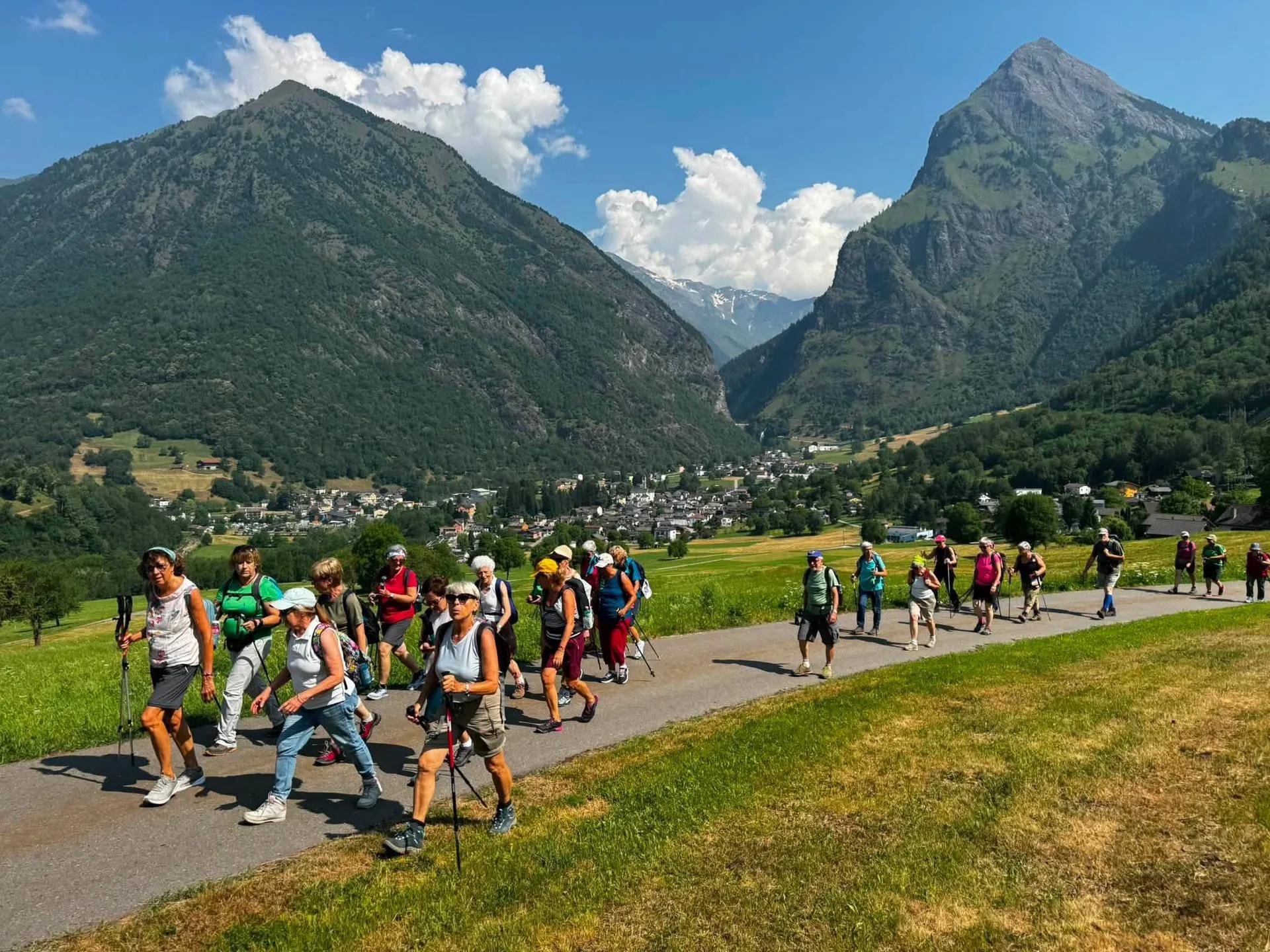 Gruppo di camminatori con bastoncini lungo una strada in una valle verde, con montagne e un paese sullo sfondo
