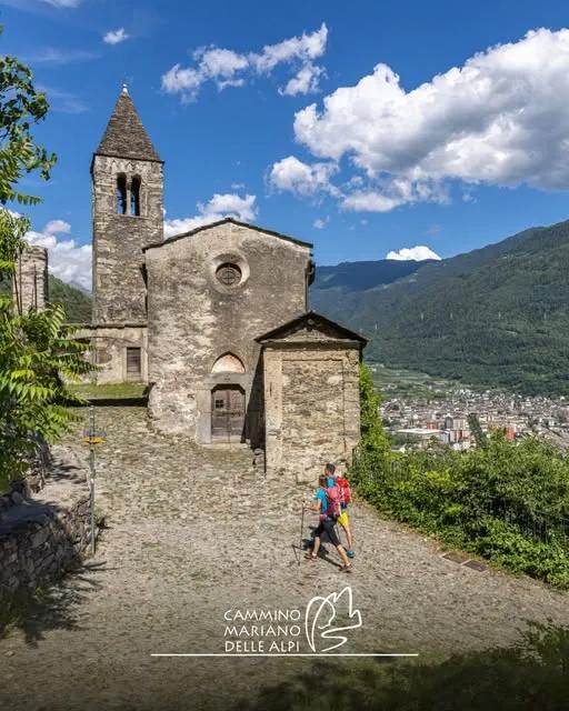 Antica chiesa in pietra con campanile affacciata sulla Valtellina, con la città in fondo alla valle e le montagne boscose, lungo il Cammino Mariano delle Alpi